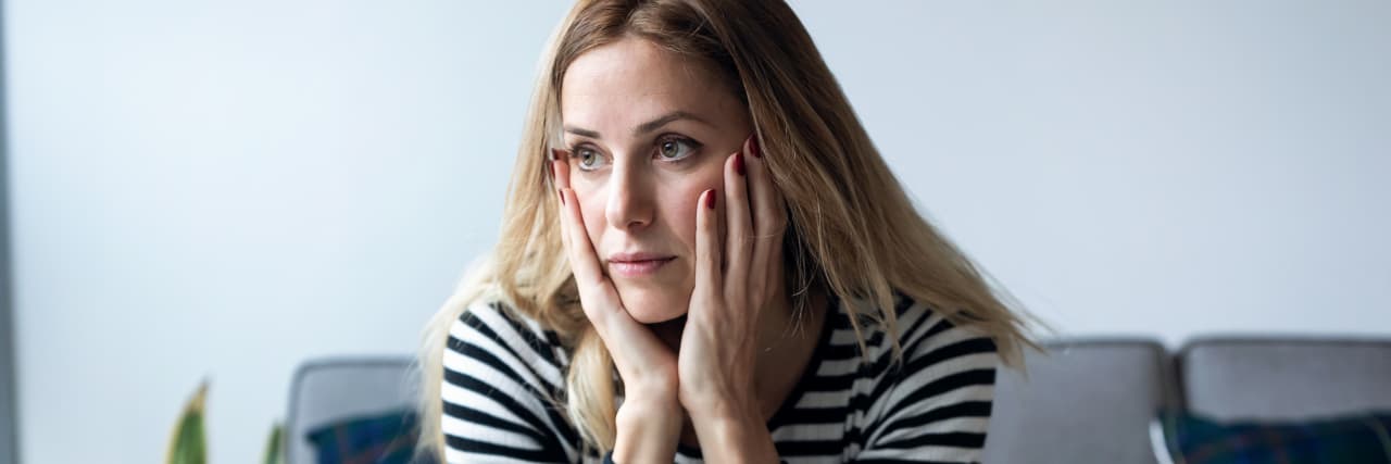 Celiac Disease and Adaptive Stress During the COVID-19 Pandemic A woman with blonde hair wearing a striped shirt and jeans sits on a couch with her hands on her face.