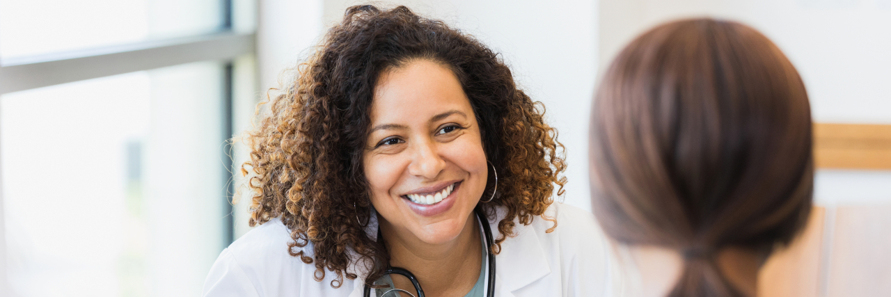 A woman with brown hair in a ponytail wearing a gray sweater talks with a female doctor of color with curly hair.