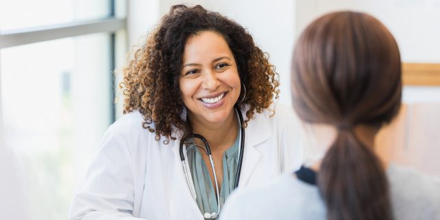 A woman with brown hair in a ponytail wearing a gray sweater talks with a female doctor of color with curly hair.