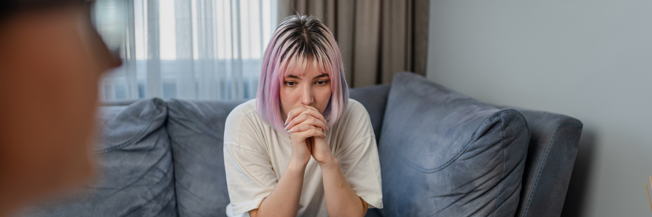 Asking a Therapist for Help With Going Back to Mental Health Treatment A woman with purple hair wearing a white shirt and jeans stares ahead as her therapist takes notes.