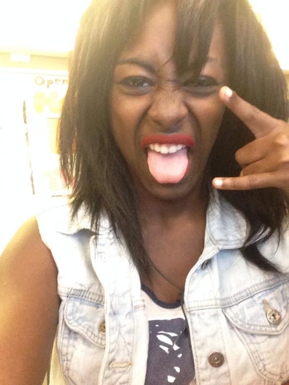 A girl throwing up punk rock hand signs with red lipstick and her tongue showing in a denim jacket and tank top