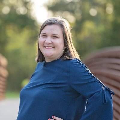 A woman with a brown bob and wearing a blue shirt smiles with her hand on her hip.