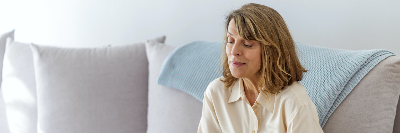 The Guilt I’m Experiencing While Grieving the Loss of My Husband Woman sitting on gray sofa, looking down with her arms wrapped around herself
