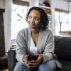 Black woman holding phone on sofa at home, smiling slightly and looking to the side
