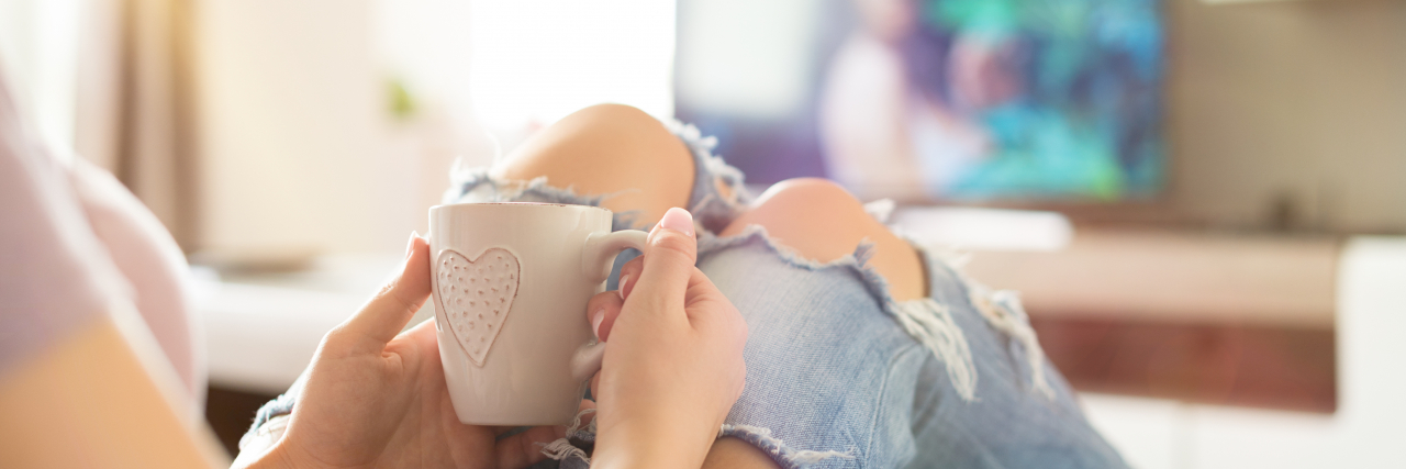 How TV Makes the World More Accessible to People With Chronic Illness Woman watching TV and drinking tea.