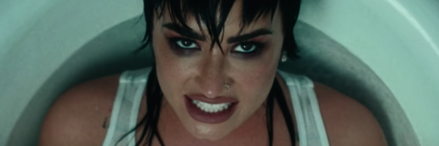 Demi Lovato sitting in a bathtub wearing a white tank top with dark makeup on her face