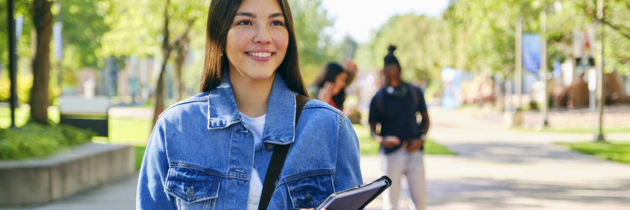 Coping With Multiple Tumors and Staying Positive A woman with long brown hair wearing a denim jacket and black pants carries a notebook and a backpack while smiling.