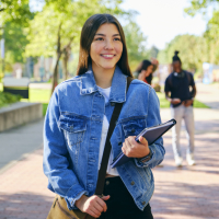 A woman with long brown hair wearing a denim jacket and black pants carries a notebook and a backpack while smiling.