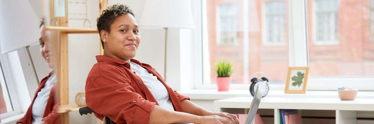 A black woman with short hair and a red shirt sits in a wheelchair while typing on a laptop and smiling confidently.