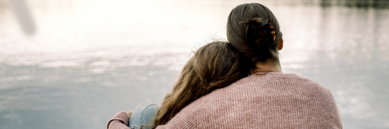 A woman with brown hair in a bun and wearing a sweater hugs a teenage girl with long, wavy hair.