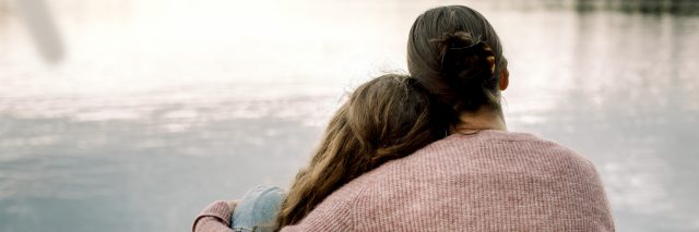 A woman with brown hair in a bun and wearing a sweater hugs a teenage girl with long, wavy hair.