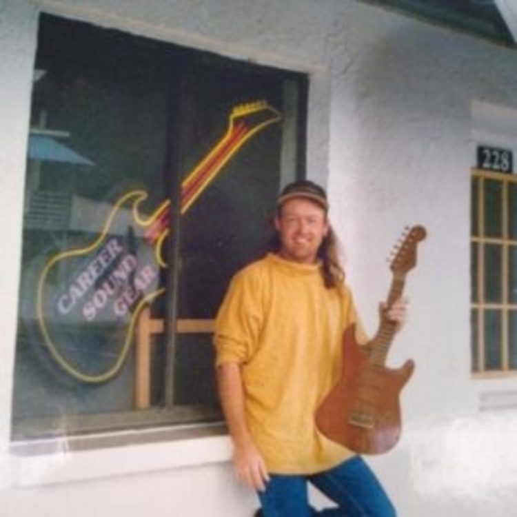 The author's friend, a man with long, red hair holds a guitar while standing next to a guitar display. 