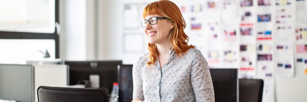 A woman with red hair wearing a collared shirt and jeans and holding a coffee mug smiles in an office break room.
