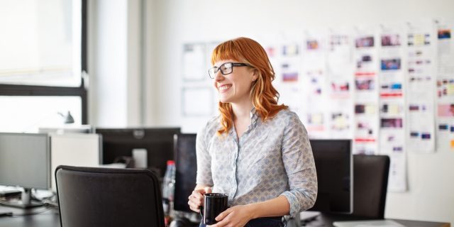 A woman with red hair wearing a collared shirt and jeans and holding a coffee mug smiles in an office break room.