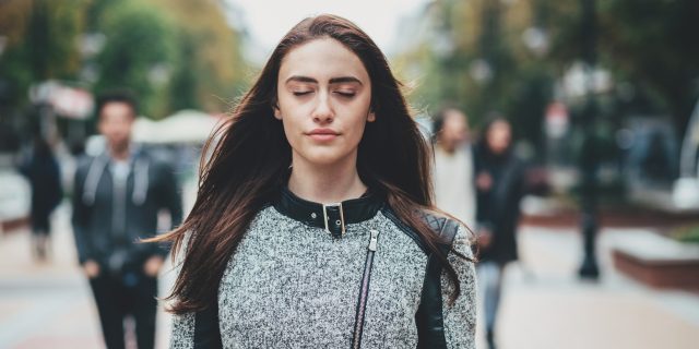 A woman with long brown hair wearing a gray coat stands and breathes with her eyes closed.