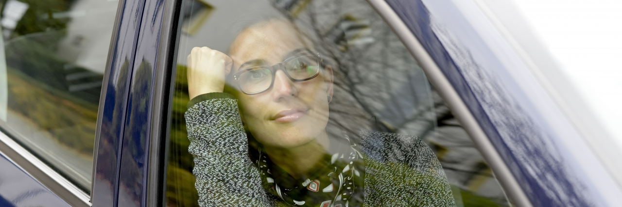 A woman with browns hair wearing a thick sweater and glasses looks out the passenger-side window of a blue car.