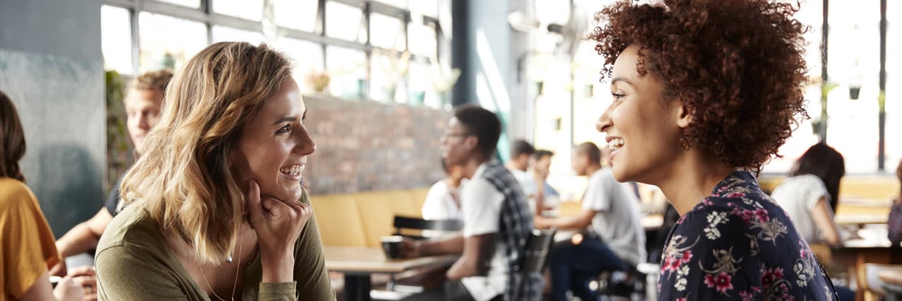 How to Set Healthy Boundaries to Improve Your Mental Health A blonde woman wearing an olive-colored shirt and a woman of color wearing a floral top talk with each other in a coffee shop.