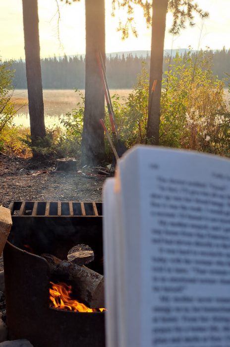 Photo of page of book in front of campfire, water and trees 
