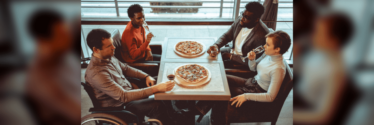 Four friends gathered at a restaurant making celebratory toast with raising glasses up and eating pizzas. One of them is disabled and is sitting in a wheelchair.