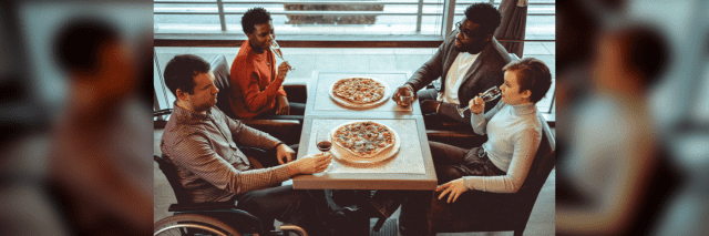 Four friends gathered at a restaurant making celebratory toast with raising glasses up and eating pizzas. One of them is disabled and is sitting in a wheelchair.