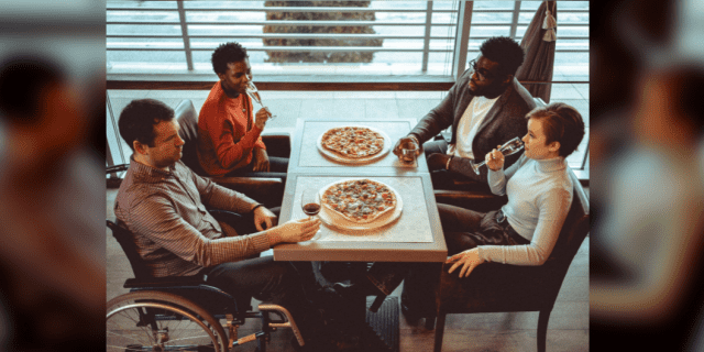 Four friends gathered at a restaurant making celebratory toast with raising glasses up and eating pizzas. One of them is disabled and is sitting in a wheelchair.