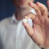 Man's hand holding a single round pill, with his face blurred in the background