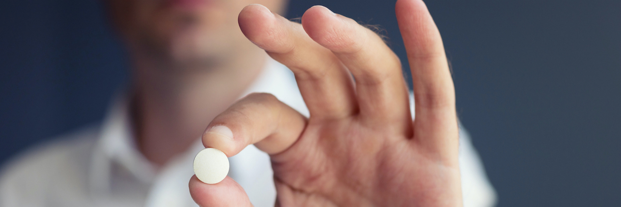 Man's hand holding a single round pill, with his face blurred in the background