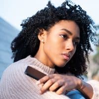Black woman using mobile phone staring forlornly into the distance in a sweater outside.