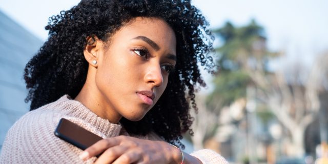 Black woman using mobile phone staring forlornly into the distance in a sweater outside.