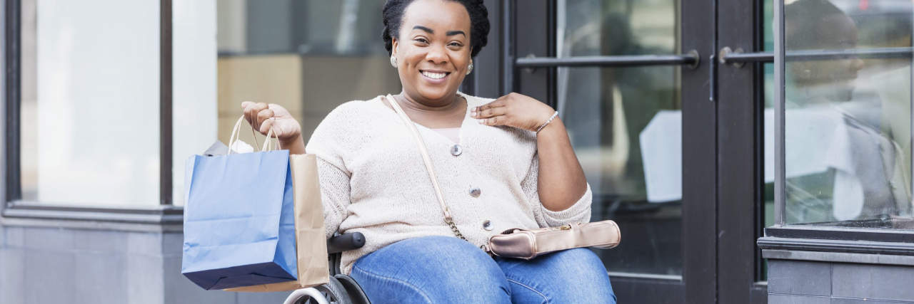 Why Companies Need to Embrace Disability Pride Month African-American woman in her 30s in a wheelchair, outdoors in the city, shopping. She is carrying shopping bags, smiling at the camera. She has spina bifida.