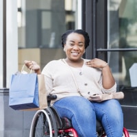 African-American woman in her 30s in a wheelchair, outdoors in the city, shopping. She is carrying shopping bags, smiling at the camera. She has spina bifida.