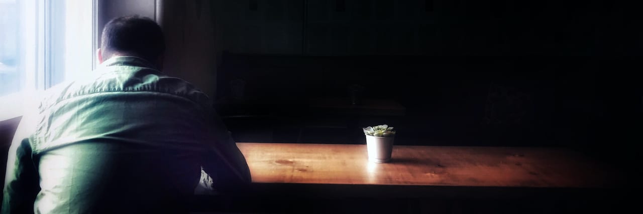 Being Told I Was Feminine and Not Man Enough Affected My Mental Health Photo of man sitting at table in the dark with his head facing downwards and a small plant on the table bathed in light from the window