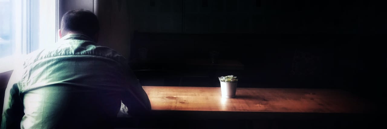 Photo of man sitting at table in the dark with his head facing downwards and a small plant on the table bathed in light from the window