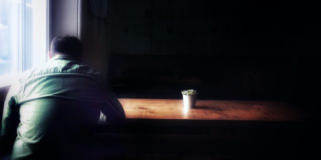 Photo of man sitting at table in the dark with his head facing downwards and a small plant on the table bathed in light from the window