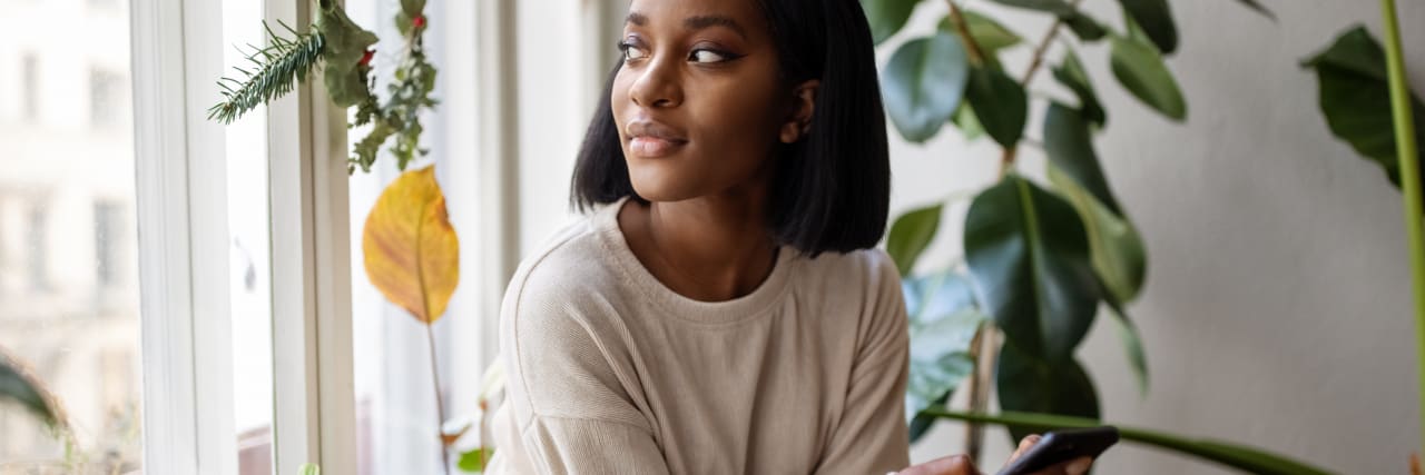 Should You Live Alone When You're Mentally Ill? Woman sitting on the windowsill with mobile phone and looking away. Woman relaxing at home looking outside the window.