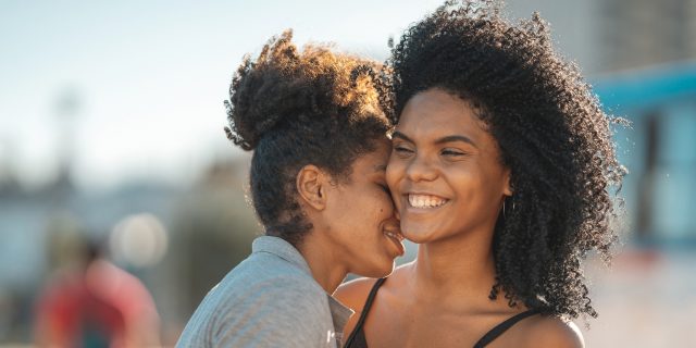 Two young women where one is kissing the other's neck. It's daylight out and one is in a cut off shirt and the other is in a Black top. They're very happy and in love!