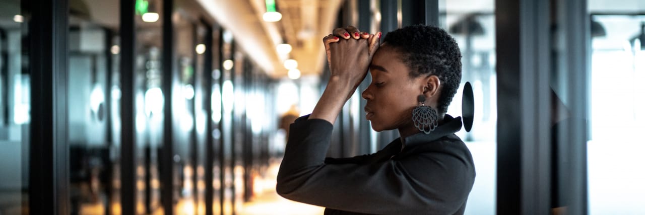 A distressed young business woman stands in an office corridor. Her hands are clasped together and raised to her forehead as she struggles with her emotions.