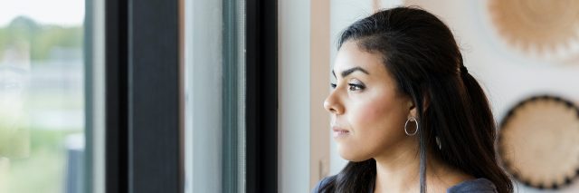 Woman of color stands holding a mug and looking out window