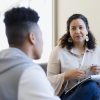 Counselor, a woman of color, listens and holds a notebook during a session with a young man