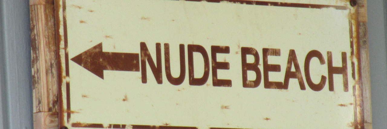 A metal sign attached to a piece of wood hung up on gray wall that says written in brown letters with an arrow, “NUDE BEACH” on a beige background.