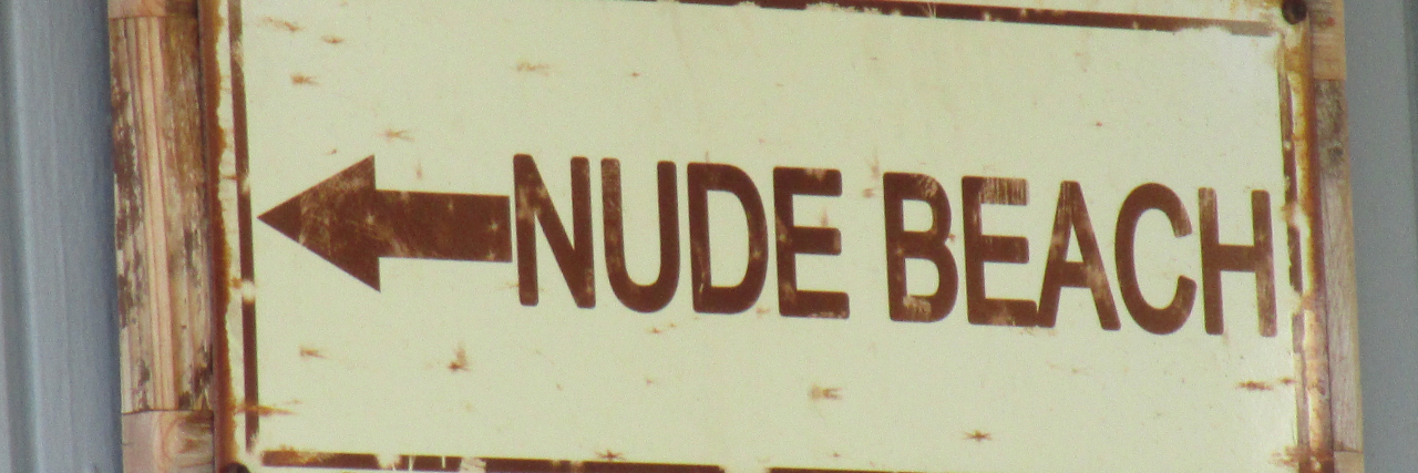 A Nude Beach Was the Best Vacation for One Woman With Body Dysmorphia A metal sign attached to a piece of wood hung up on gray wall that says written in brown letters with an arrow, “NUDE BEACH” on a beige background.