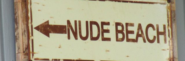A metal sign attached to a piece of wood hung up on gray wall that says written in brown letters with an arrow, “NUDE BEACH” on a beige background.