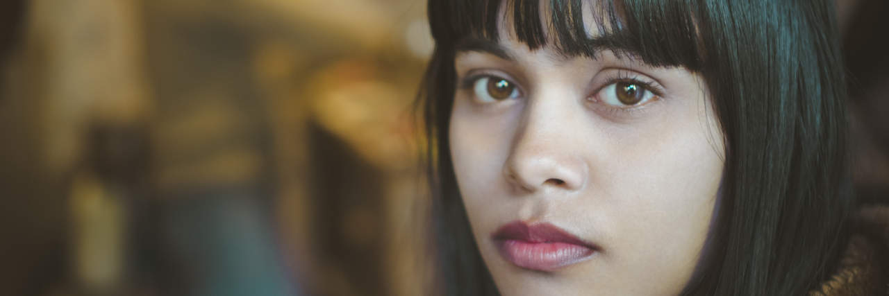 Indoor image of serene Asian, Indian young woman at home. She is sitting in kitchen at day time and looking at camera with blank expression. One person, head shot and selective focus with copy space.