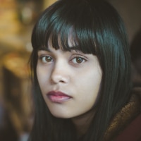 Indoor image of serene Asian, Indian young woman at home. She is sitting in kitchen at day time and looking at camera with blank expression. One person, head shot and selective focus with copy space.