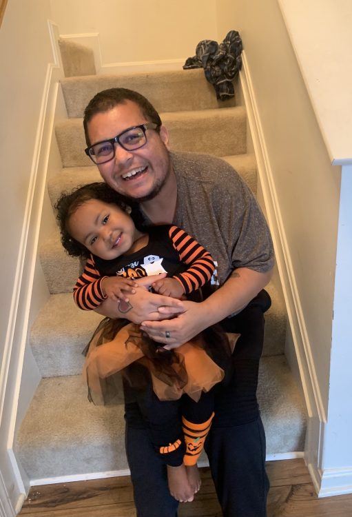 Kris, a Black transgender man holding his young daughter, smiling
