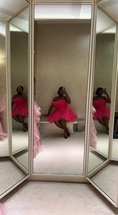 A beautiful Black woman staring in a mirror looking at herself in a big Barbie dress