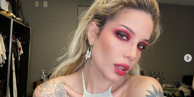 Halsey looking into the camera sporting bright eyeshadow and a lipstick in a white top with their hair up.