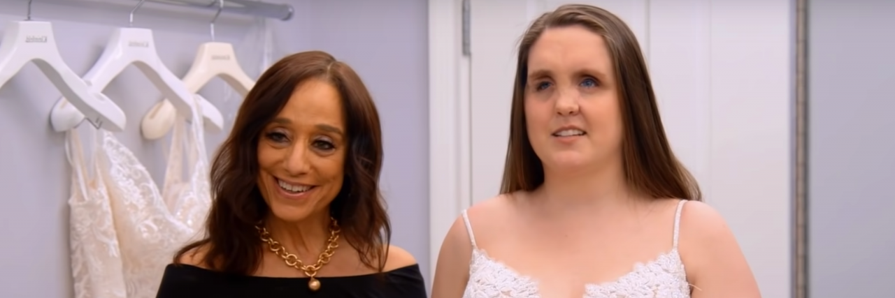 'Say Yes to the Dress' Features a Blind Bride Two women stand next to one another, one is in a bridal gown with brown hair. The gown has spaghetti straps and is plunging. They are in a dressing room with wedding dresses hanging up.