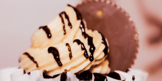 A close up shot of a chocolate cupcake with peanut butter frosting, chocolate drizzle, and a reese's cup on top.