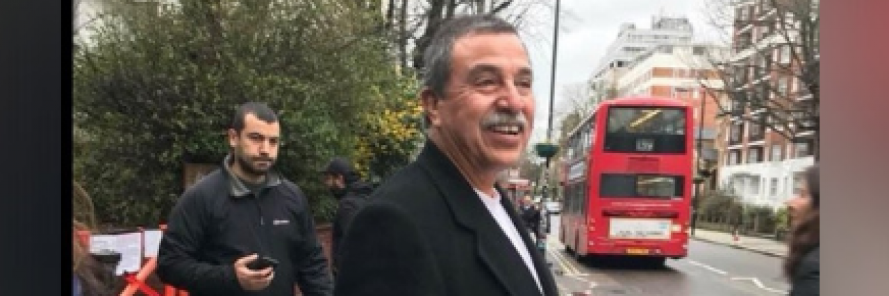 Image of Alfonso smiling and standing on sidewalk with a red double decker bus in the distance behind him